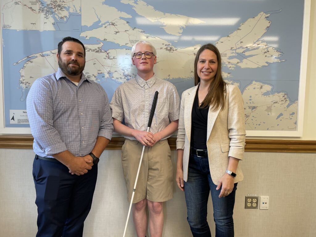 Caelin, Richard, and Minister Druhan stand in front of a map of Nova Scotia showing all public schools. 